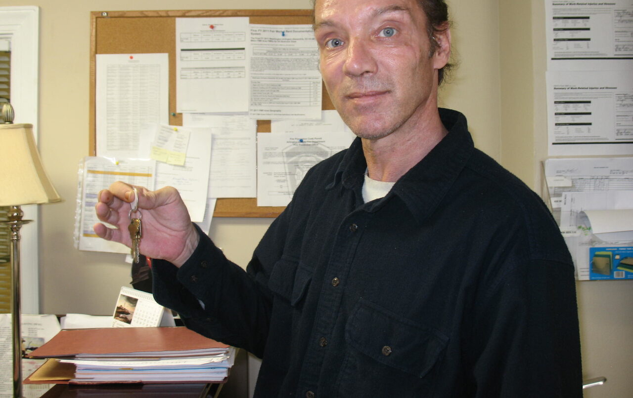 A man in a black polo holds a key while smiling for the camera