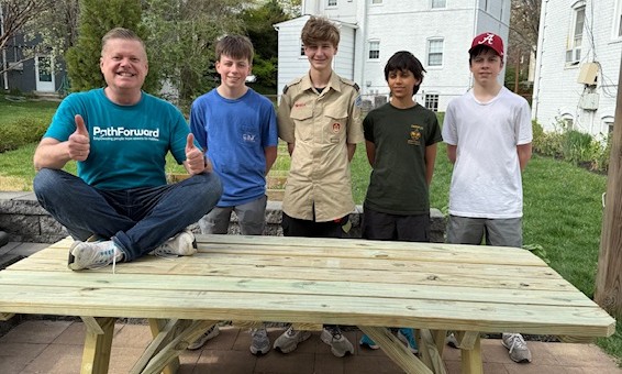 A member of the PathForward team sits on a new picnic table while smiling for the camera alongside four boys from Boy Scout Troop 162