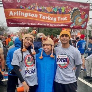 Three people wearing turkey hats stand in front of the Arlington Turkey Trot sign on a crowded street